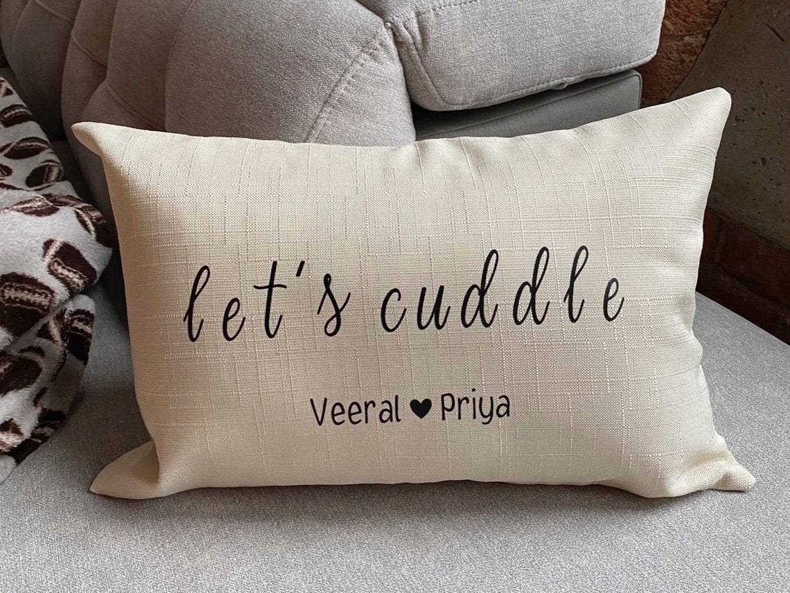 Rectangular decorative pillow with the worlds &quot;let&#x27;s cuddle&quot; and two names with a heart between them in smaller print at the bottom 