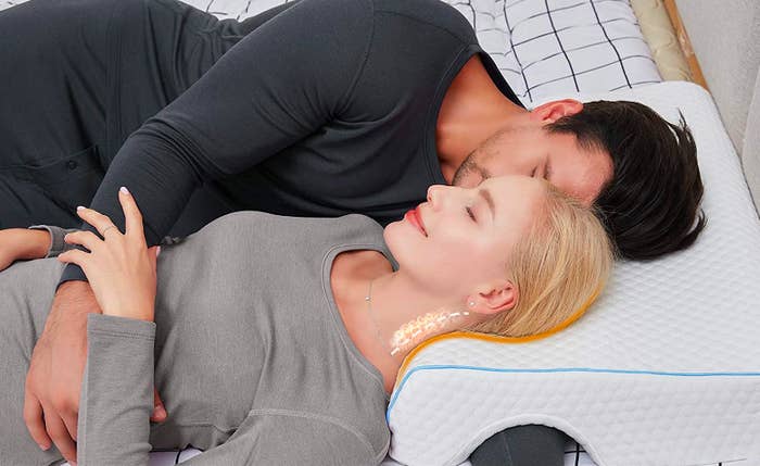 Couple snuggling in bed while both sleep on the curved memory foam pillow and one person&#x27;s arm underneath the other person, putting their arm through the hole in the bottom