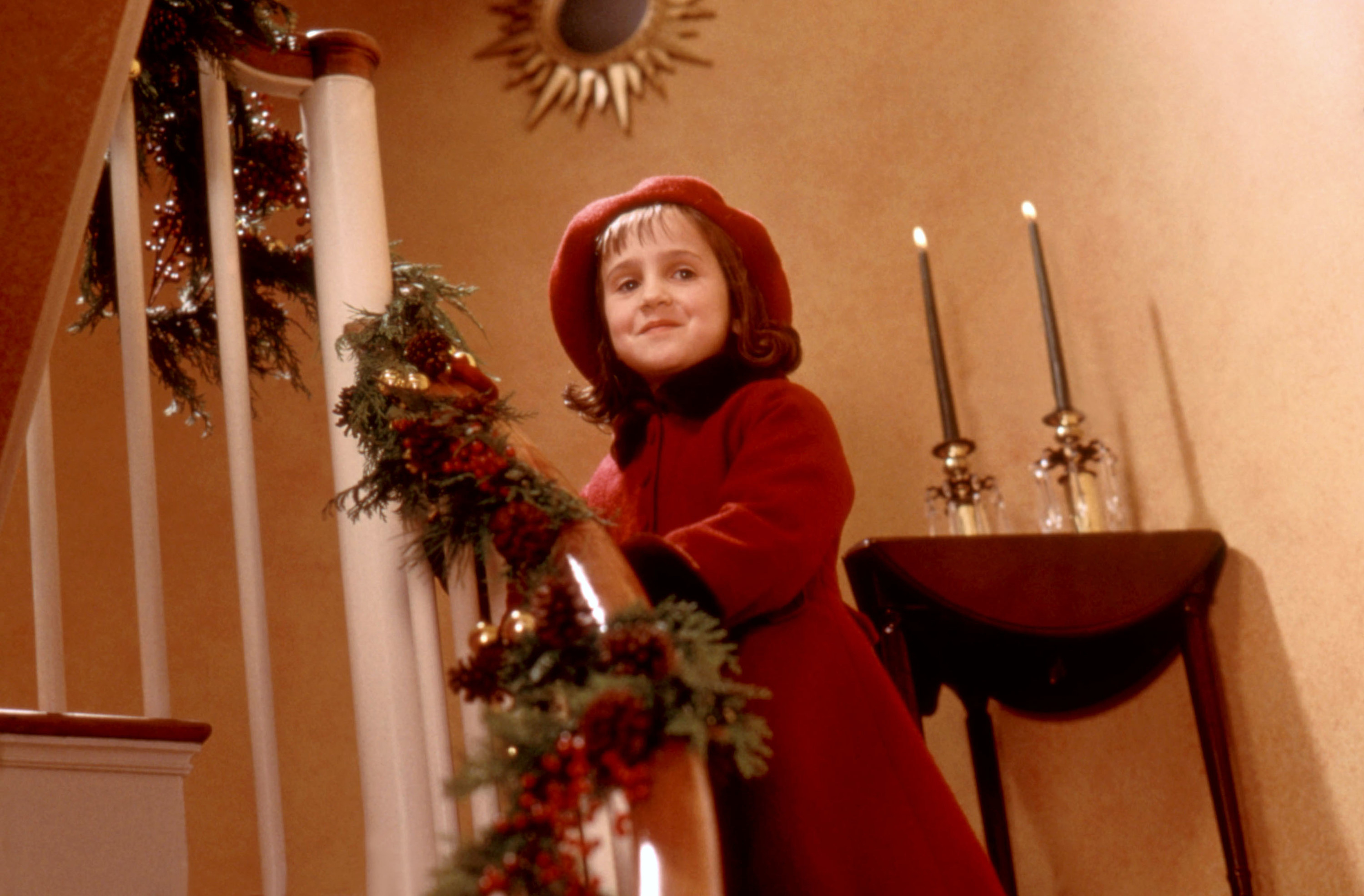 Mara Wilson standing in a staircase decorated for Christmas while wearing a winter coat and beret in Miracle on 34th Street
