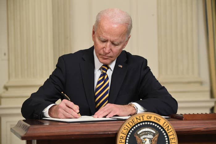 Joe Biden sits and signs a document at a desk with the presidential seal on it