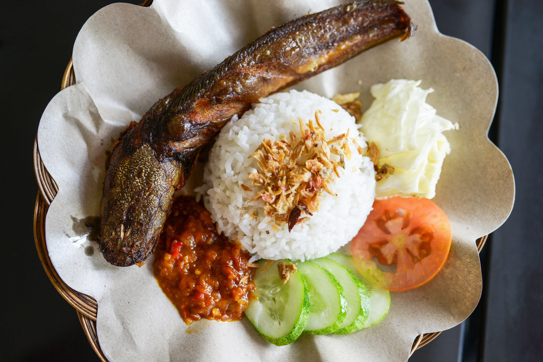 A whole fried fish sits on a plate with a mound of rice, veggies, and a spicy-looking sauce