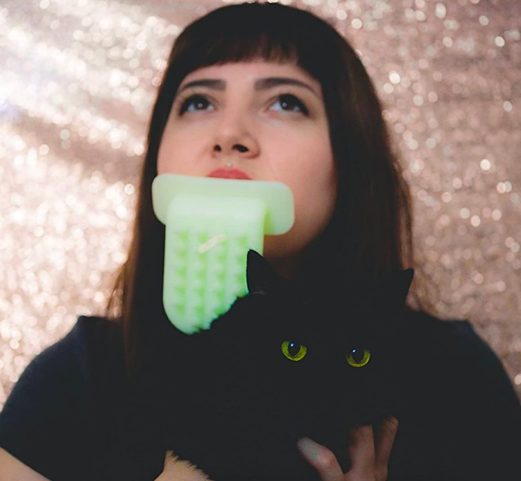 Person licking cat with a giant, silicone, tongue-shaped scratcher they are holding with their mouth 