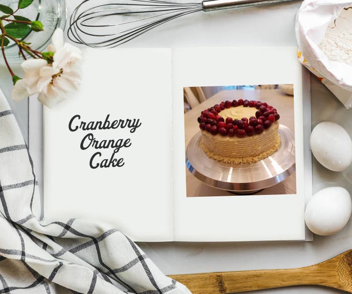 An overhead shot of cookbook on a table surrounded by ingredients and a photo on the inside of a white cake with cranberries surrounding the perimeter. 
