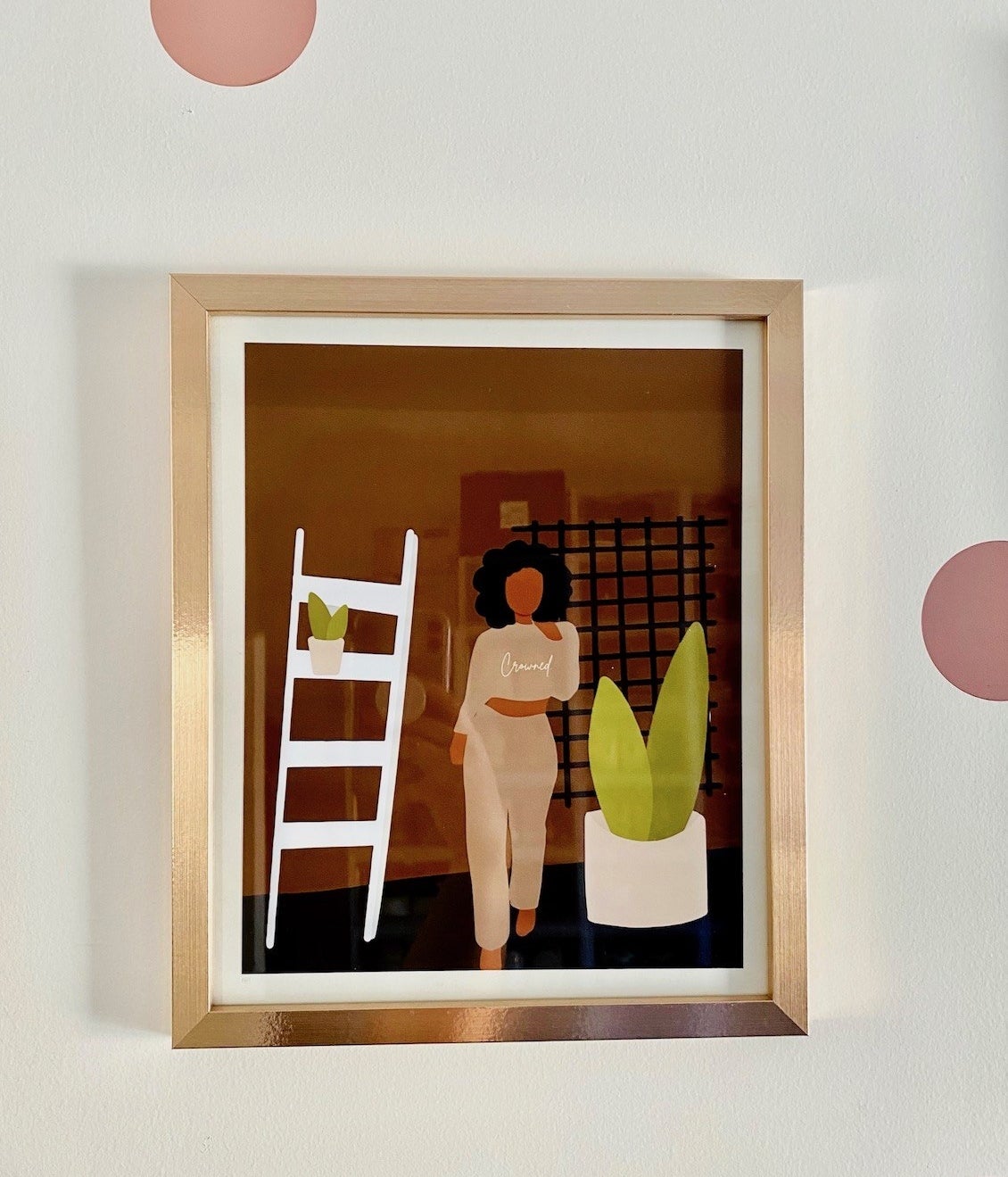 A framed print of an illustrated Black woman wearing a sweatshirt that says "crowned" while standing next to plants