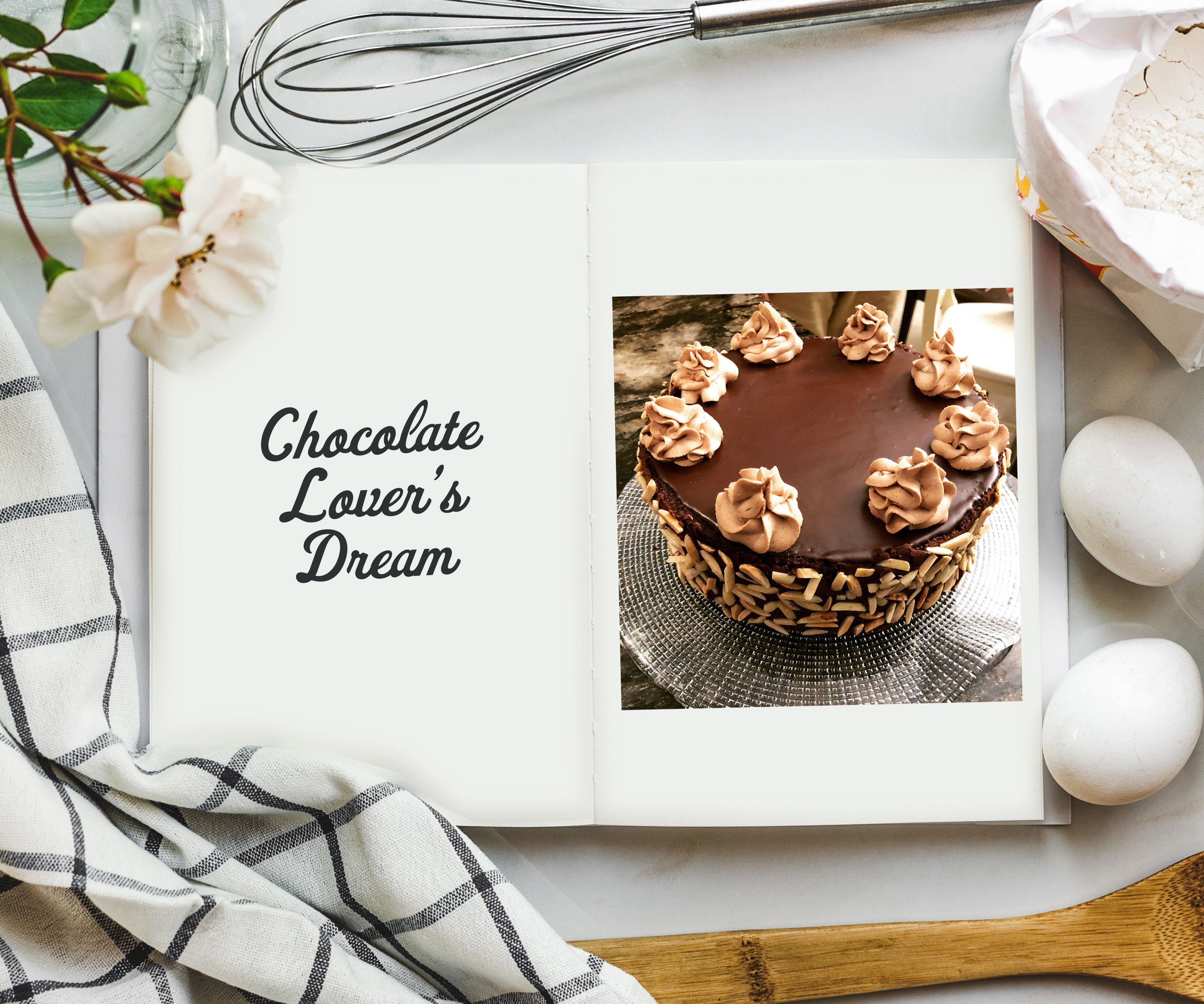 An overhead shot of cookbook on a table surrounded by ingredients and a photo on the inside of a dark chocolate cake on a cake stand with slivered almonds on the side and white piped florets on the top.