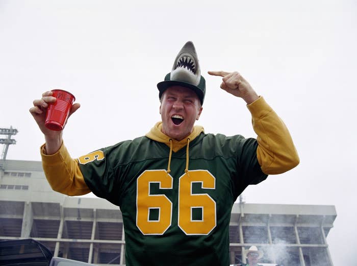 A tailgating football fan pointing at his baseball cap which has a shark head on top and holding a Solo cup