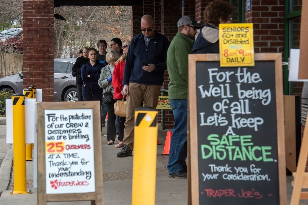 A line of customers wait to enter a Trader Joe&#x27;s store behind signs saying &quot;Let&#x27;s keep a safe distance&quot; and &quot;We are limiting 25 customers at a time&quot;