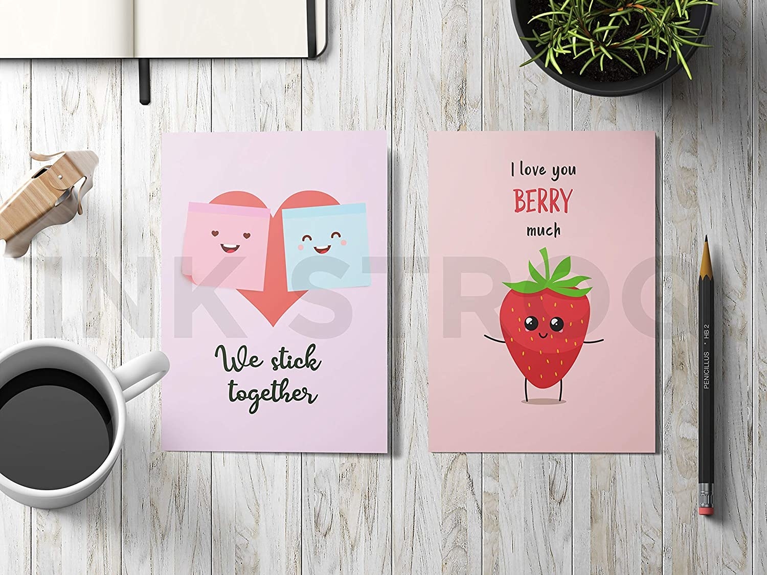 A wooden desk with a coffee mug, journal, plant, and two pink postcards on it. One has a drawing of sticky notes and says, &quot;We stick together&quot;. The other one has a drawing of a strawberry and says, &quot;I love you berry much&quot;.