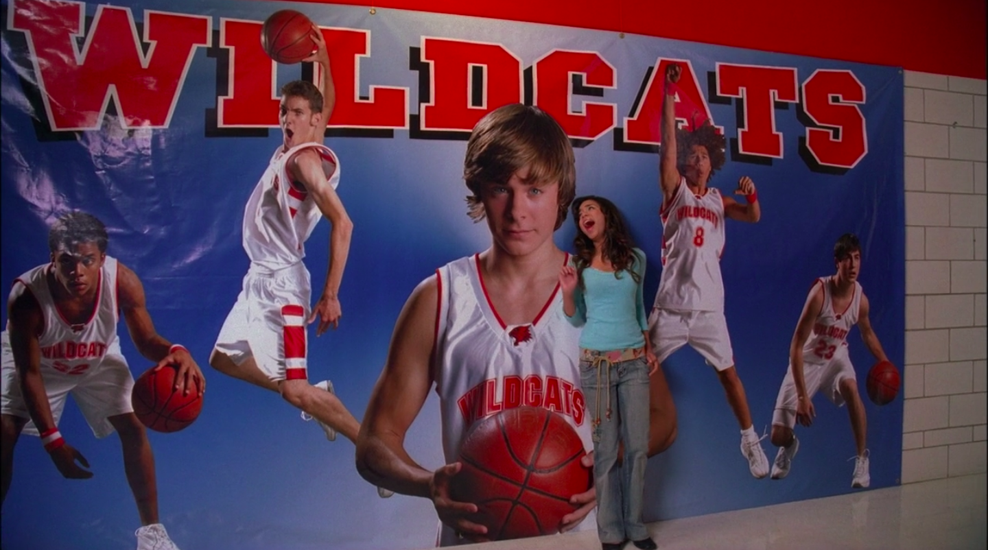 Gabriella sings in front of a poster of the basketball team with Troy front and center