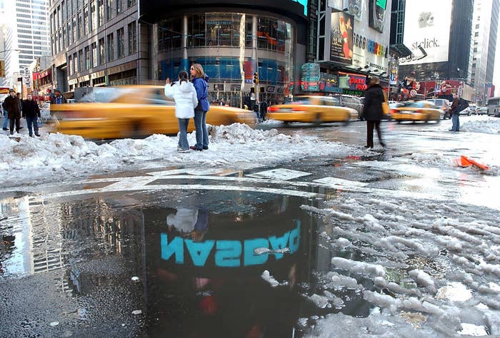 A pool of melted snow with a neon sign reflected in the dirty water as taxis drive by and pedestrians wait to cross the street