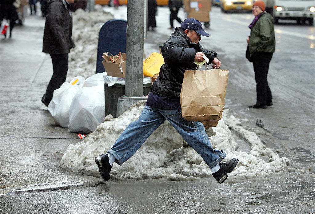 A man carrying a paper bag trues to jump across a large pool of filthy slush so he can cross the street