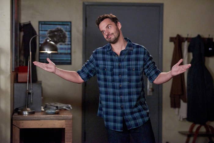 Nick Miller in a blue plaid shirt shrugging in the entryway to his apartment