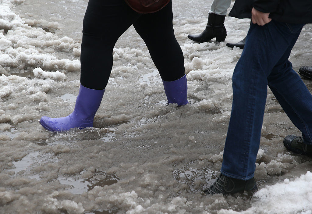 A closeup of a person wearing rain boots and another person wearing snow shoes as they walk through dirty slush