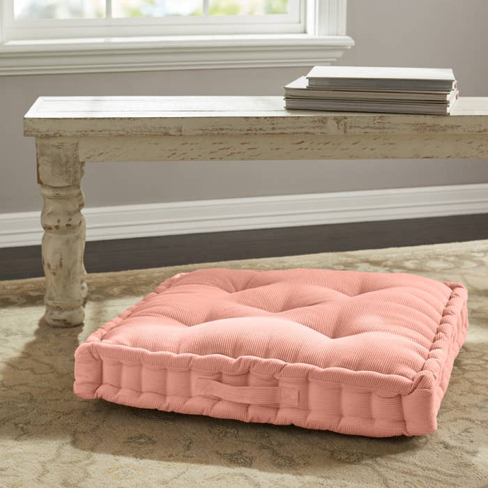The cushion, which is square, tufted, and has a small handle on the side, in dusty pink