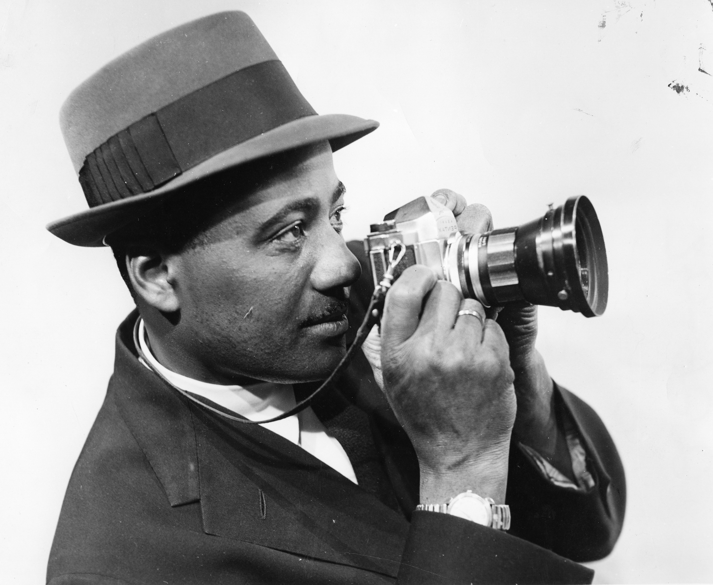 A man in a suit and hat aiming an old film camera