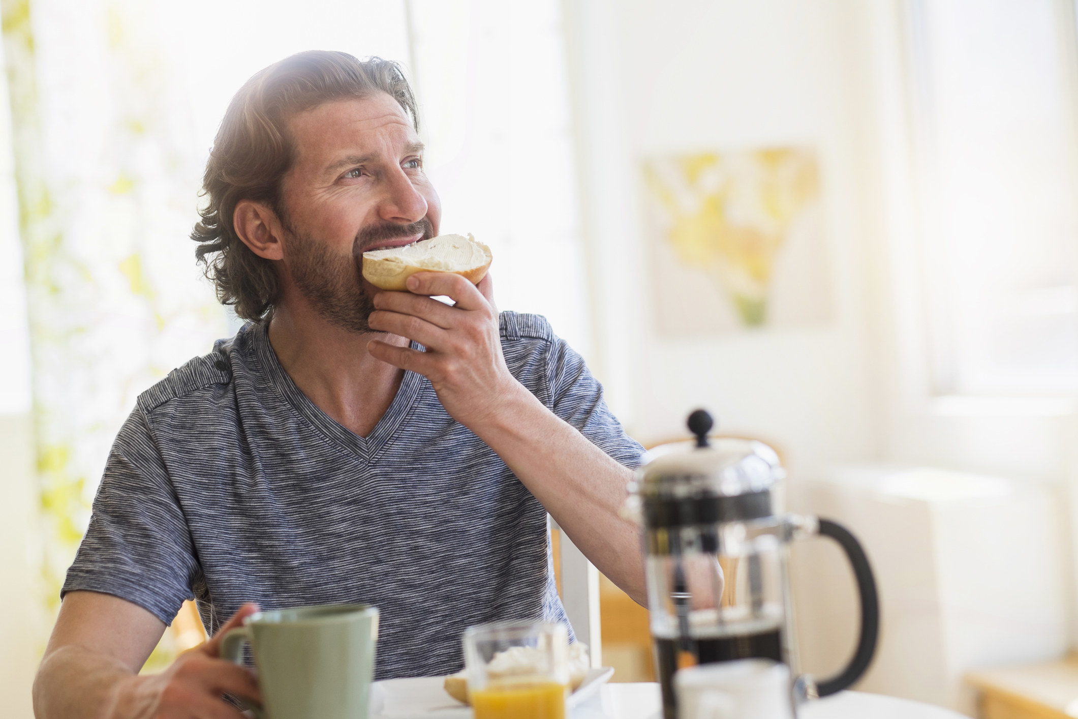 A man sitting in a white kitchen eating a bagel next to some coffee and orange juice.
