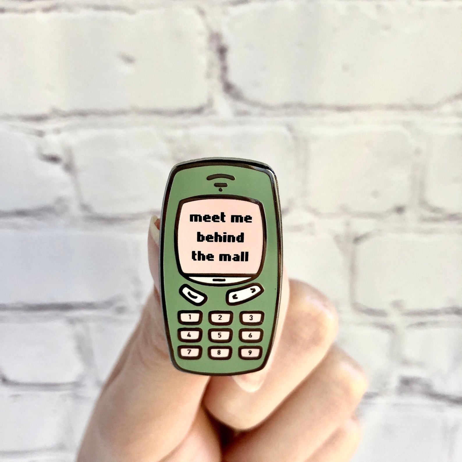 Model holding an enamel pin of a green cell phone with the text &quot;meet me behind the mall&quot; 
