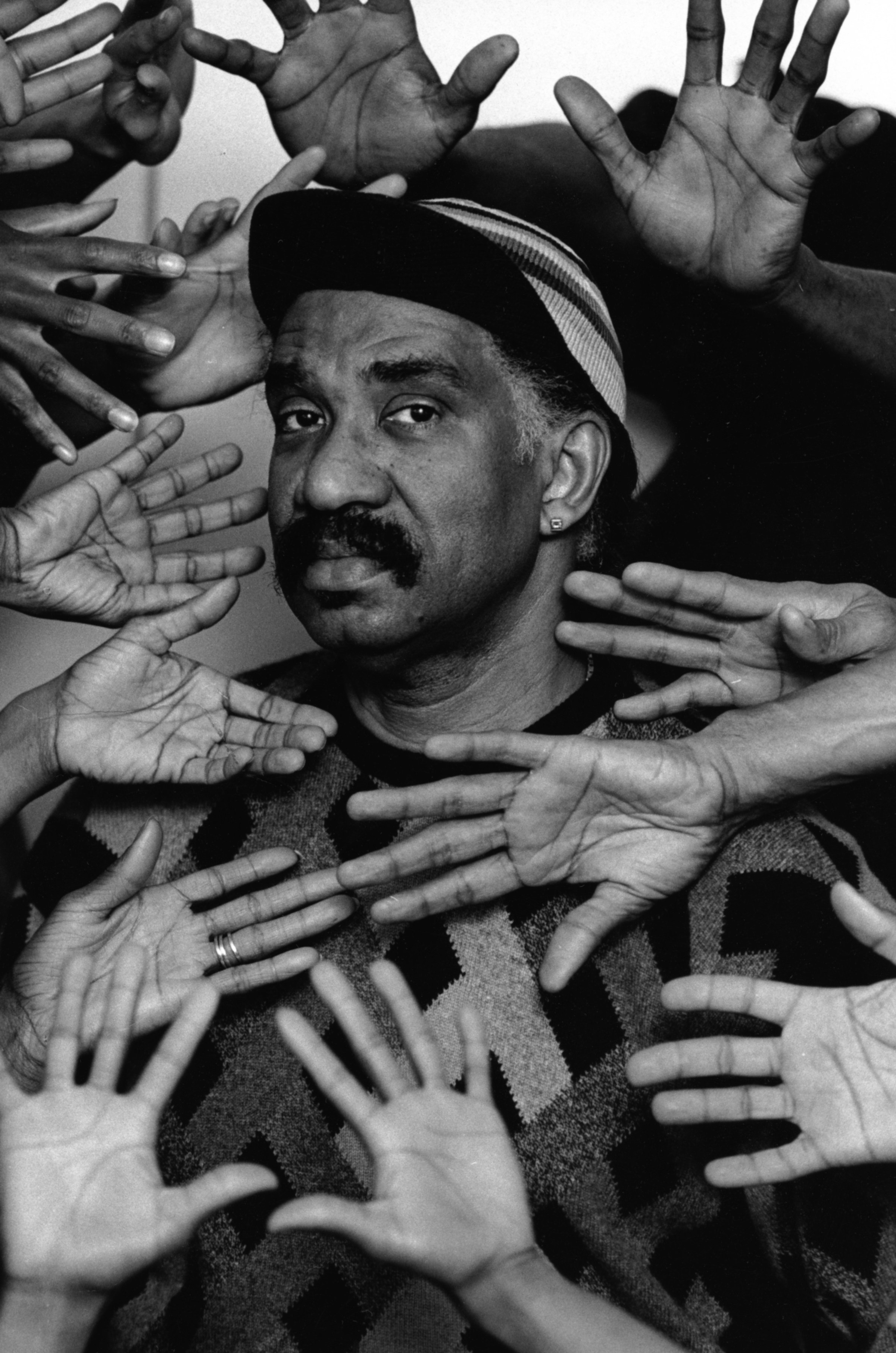A man in a hat and sweater with many people&#x27;s hands framing his face