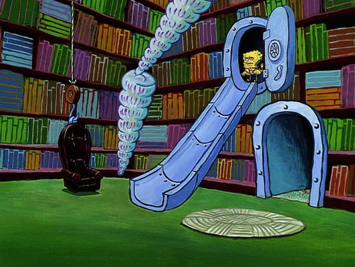 SpongeBob is standing at the top of a metal slide in a library.
