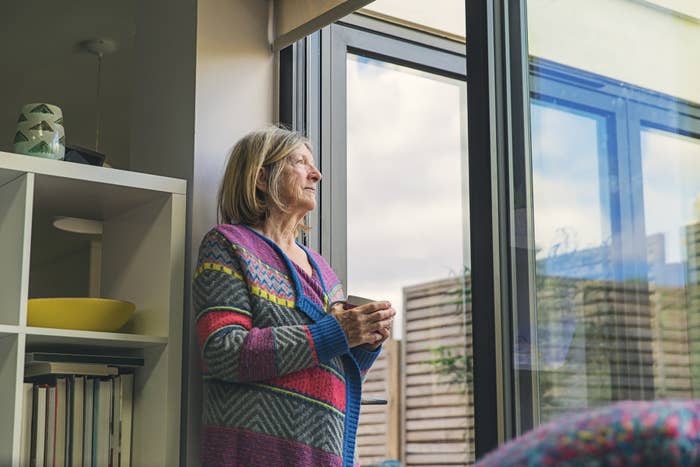 Older woman standing by her window looking out while holding a cup of coffee.