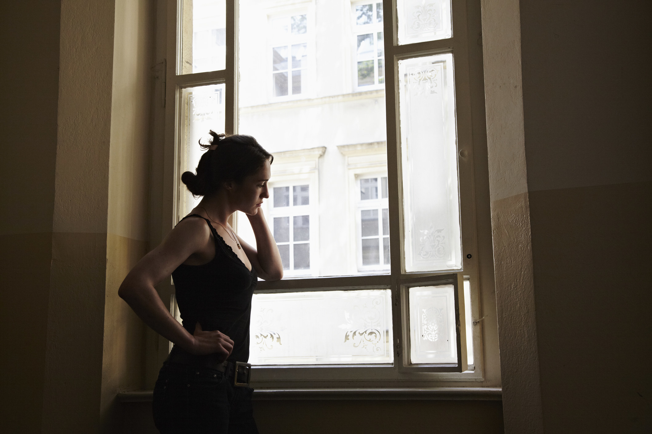 A young woman stands by a window in a dark room, deep in thought