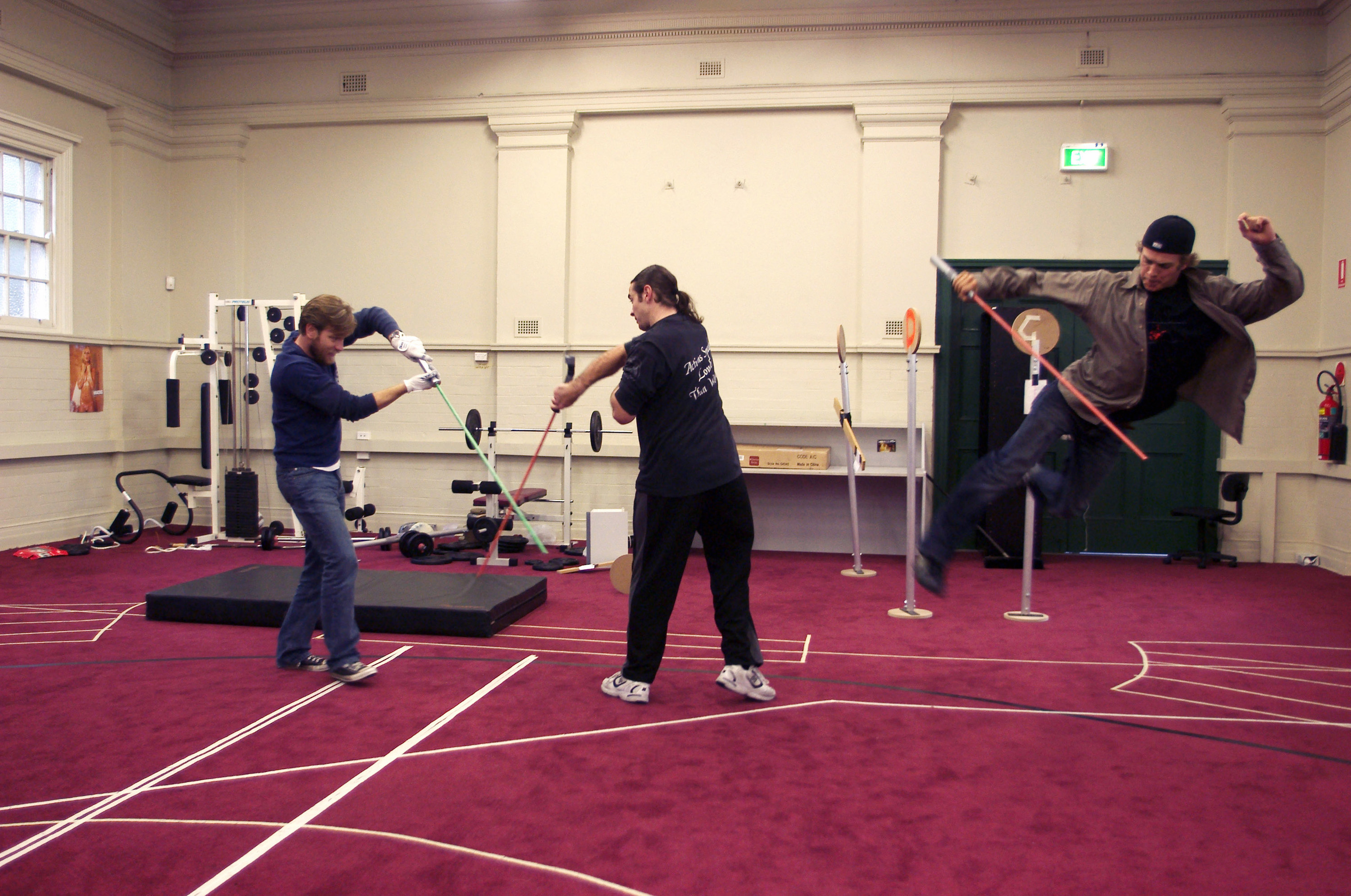 Ewan McGregor and a stunt double practicing a light saber fight with sticks in a gym as Hayden jumps and does a kick while holding a stick