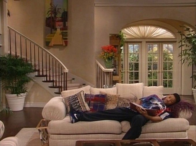 Will is laying on a white couch with colorful pillows. A curved staircase and French doors are in the background.