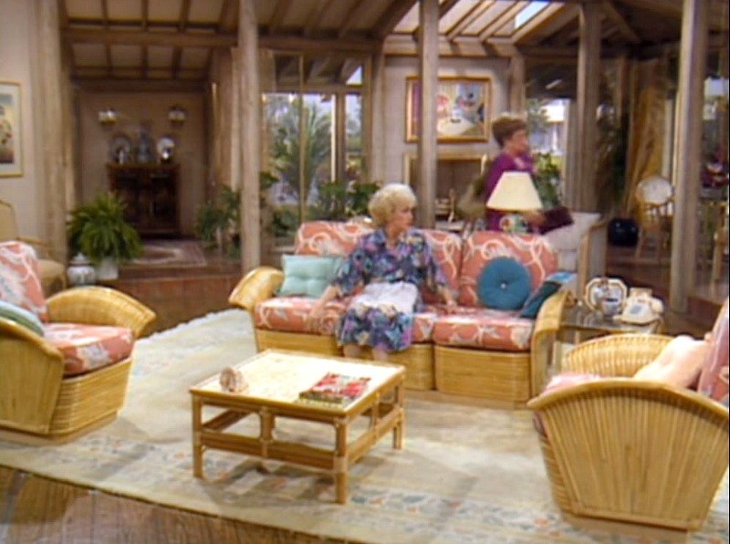 A spacious living room with wicker furniture that has pink cushions. Rose is sitting on the couch and Blanche is walking in the background.