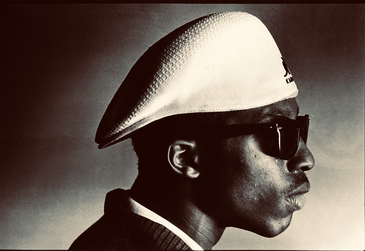 A young man in profile with sunglasses and a kangol hat on