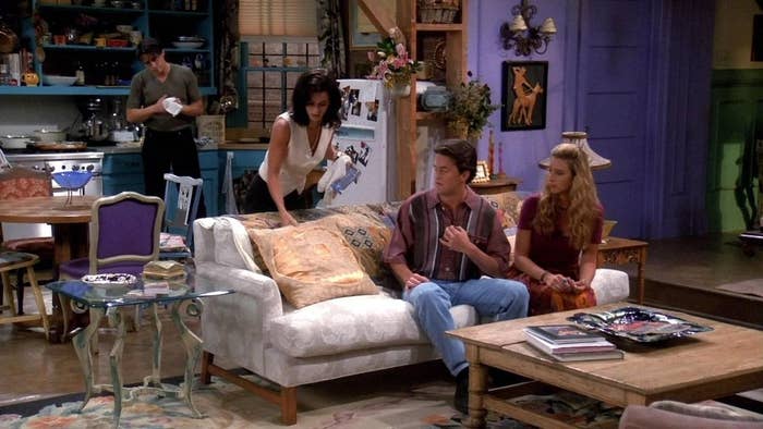 Joey in the kitchen while Monica, Phoebe, and Chandler gather around the couch in the living room