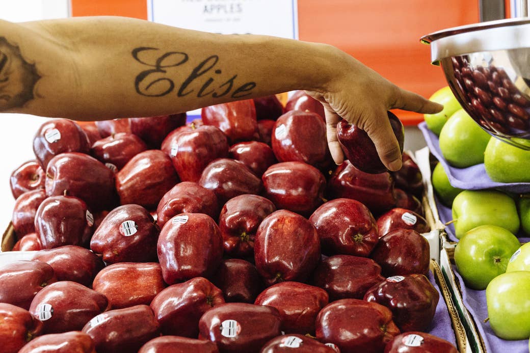 A woman&#x27;s arm, tattooed with the name &quot;Elise,&quot; reaches over a pile of apples at the supermarket