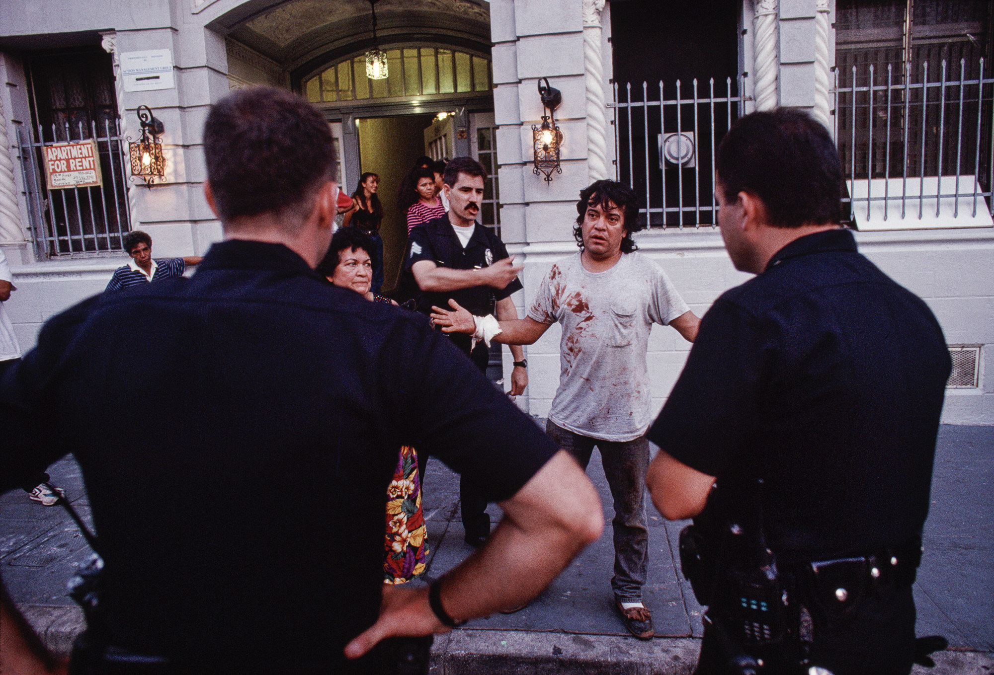 Two cops seen from behind talking to a crowd of neighbors including a man with blood on his shirt and a bandage on his wrist who looks annoyed