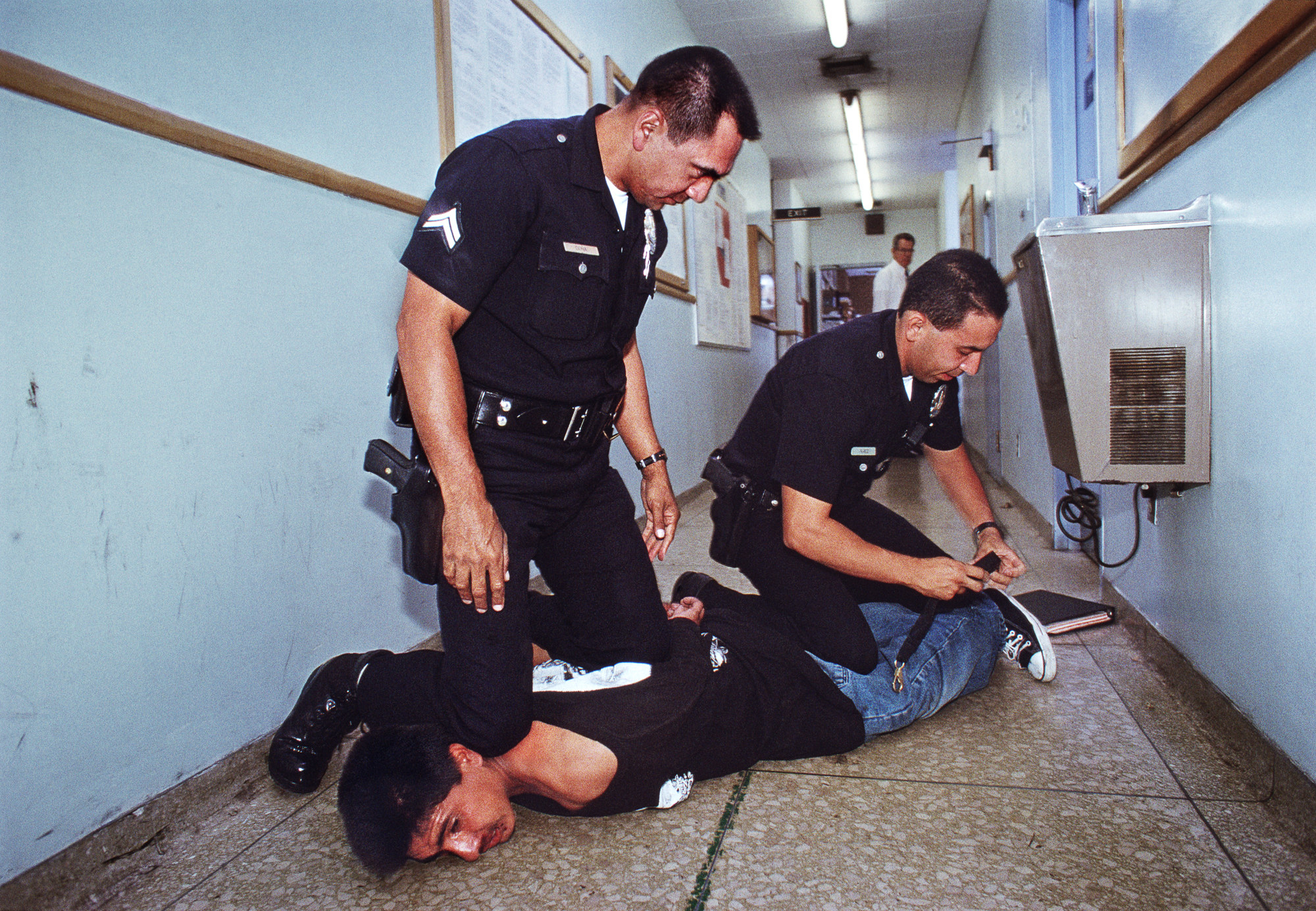 Two cops kneel on a guy in a hallway while adjusting some kind of strap