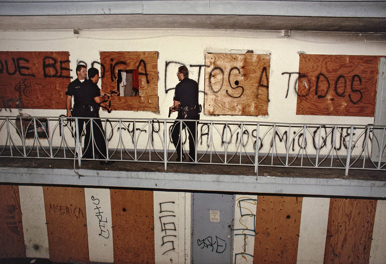 Two cops shine their flashlight into a hole on the second story of a motel that is covered in graffiti while a third cop draws his gun