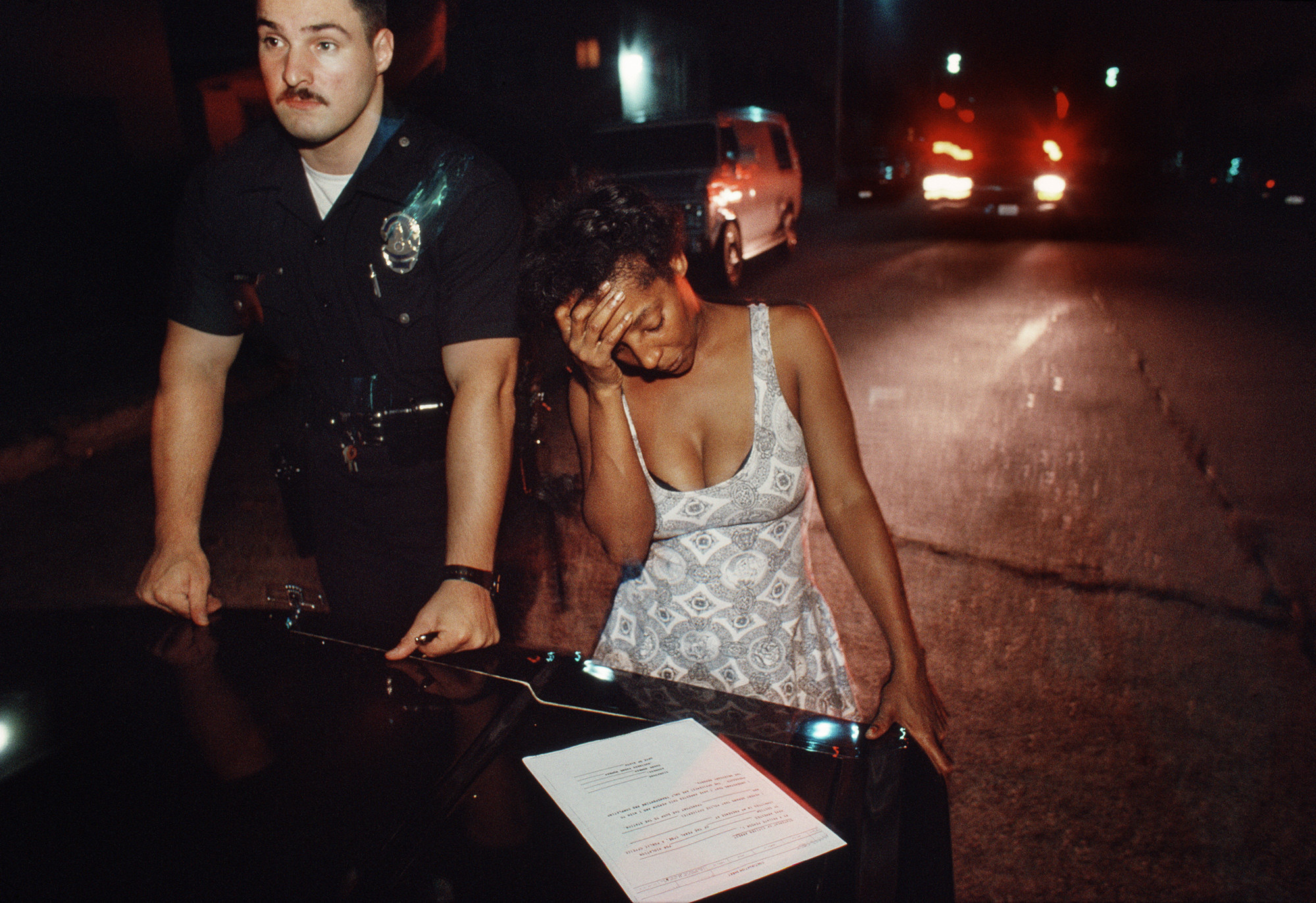 A woman holds her head in her hand while looking at a piece of paper on the trunk of a car while a police officer stands next to her and looks off into the distance