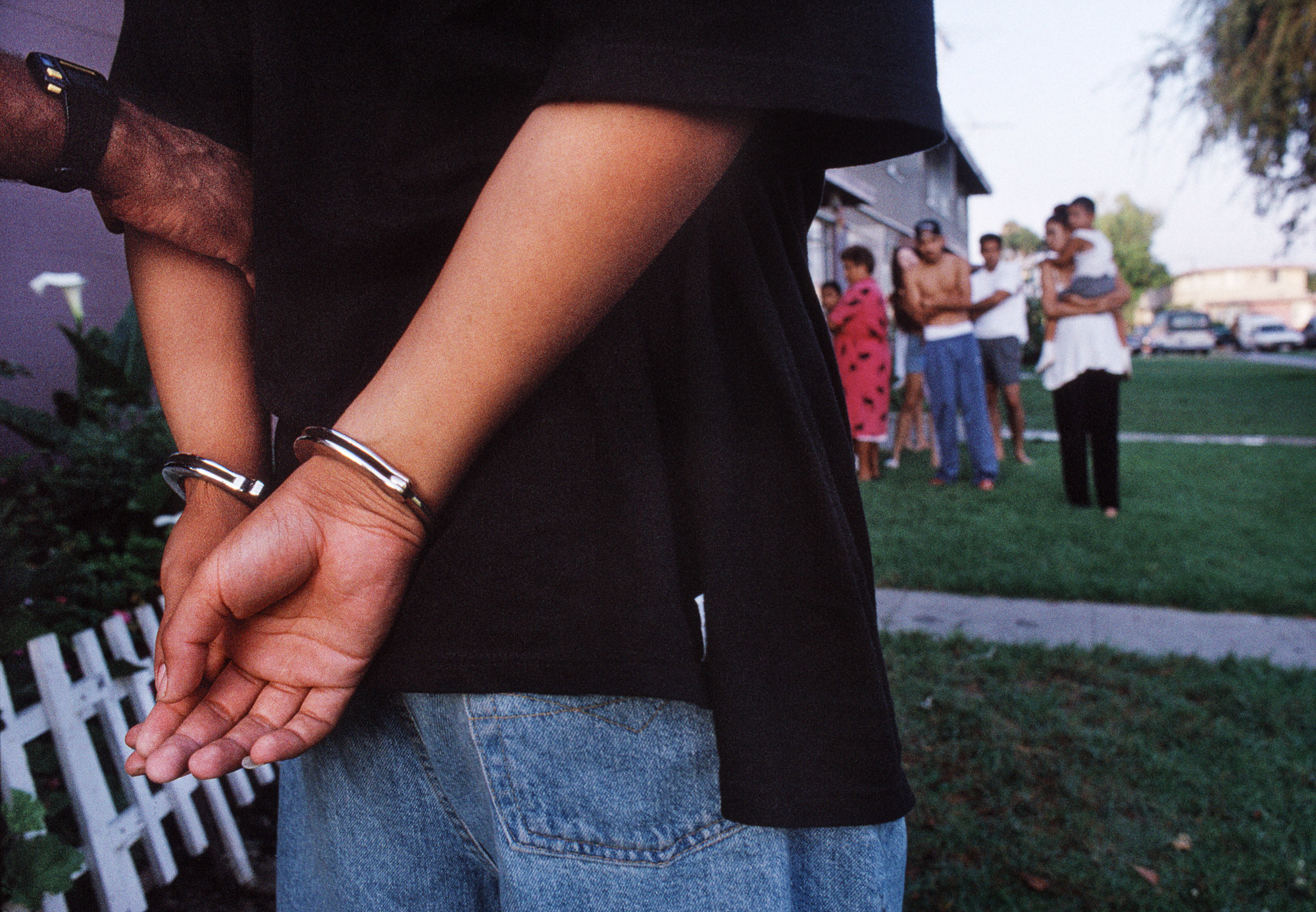 A man in handcuffs is seen from behind while a family watches in the distance, out of focus