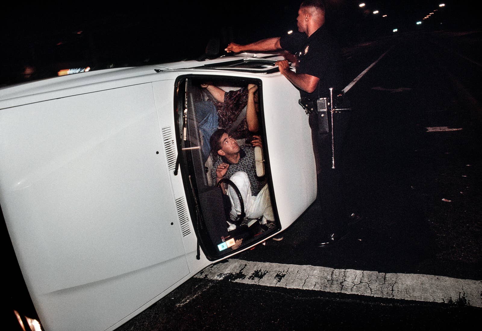 Two men are seen through the windshield of a car, which is laying on its side while a police officer tries to open the door