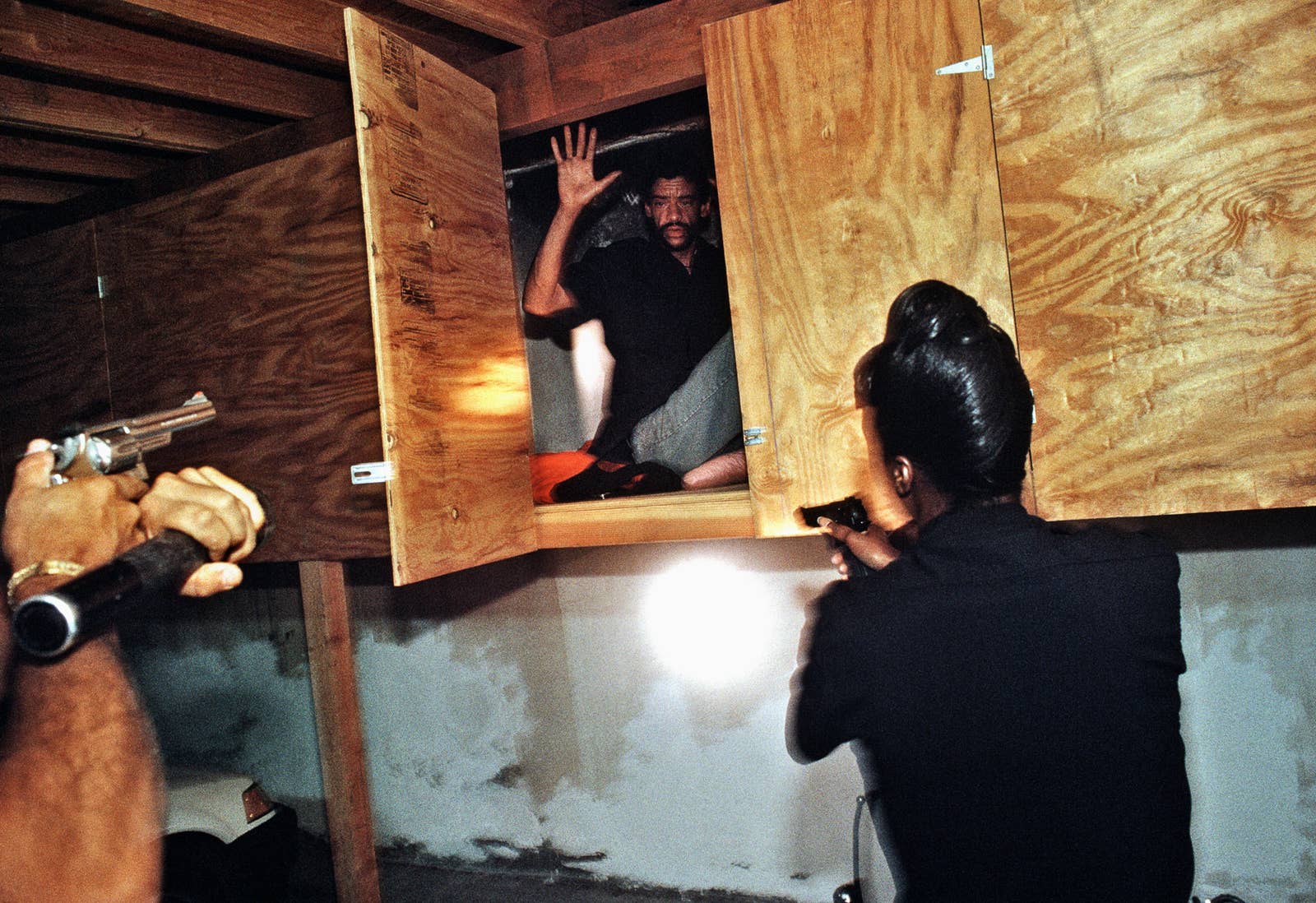 A man seen in a plywood cupboard with his hands up as two officers are seen from behind approaching with guns