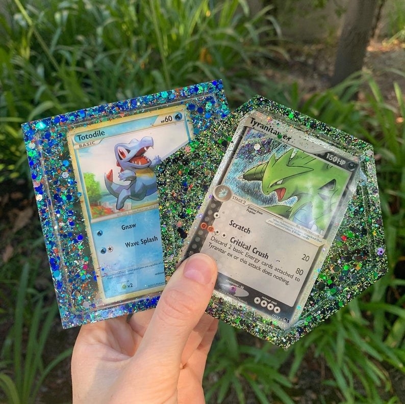Hands hold clear glitter-filled coasters with Totodile and Tyranitar Pokémon
