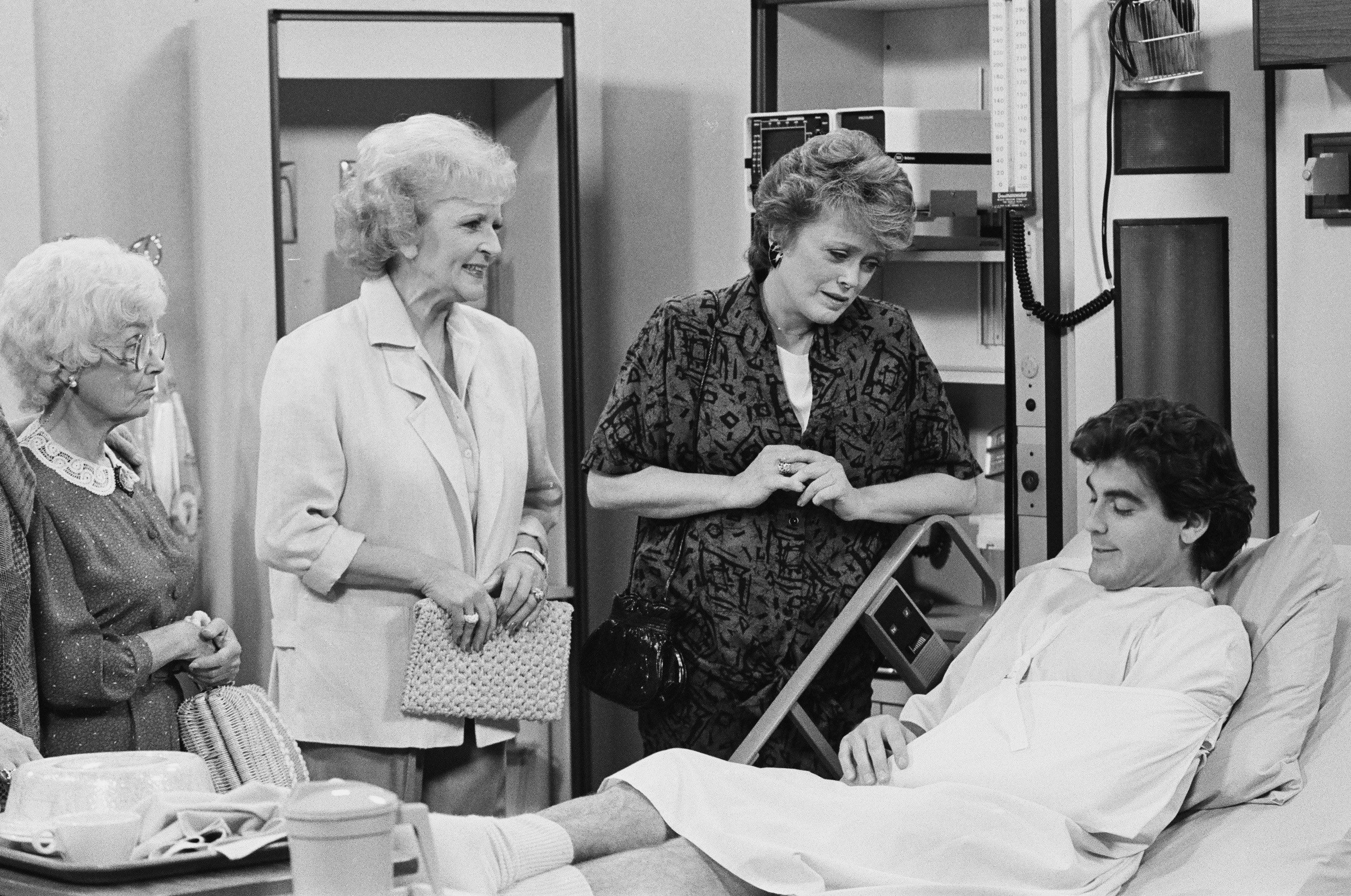 George Clooney laying in a hospital bed surround by Estelle Getty, Betty White, and Rue McClanahan 