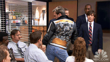 Andy Bernard from The Office dressed as George Michael and dancing in the Dunder Mifflin conference room while his co-workers look on nonplussed