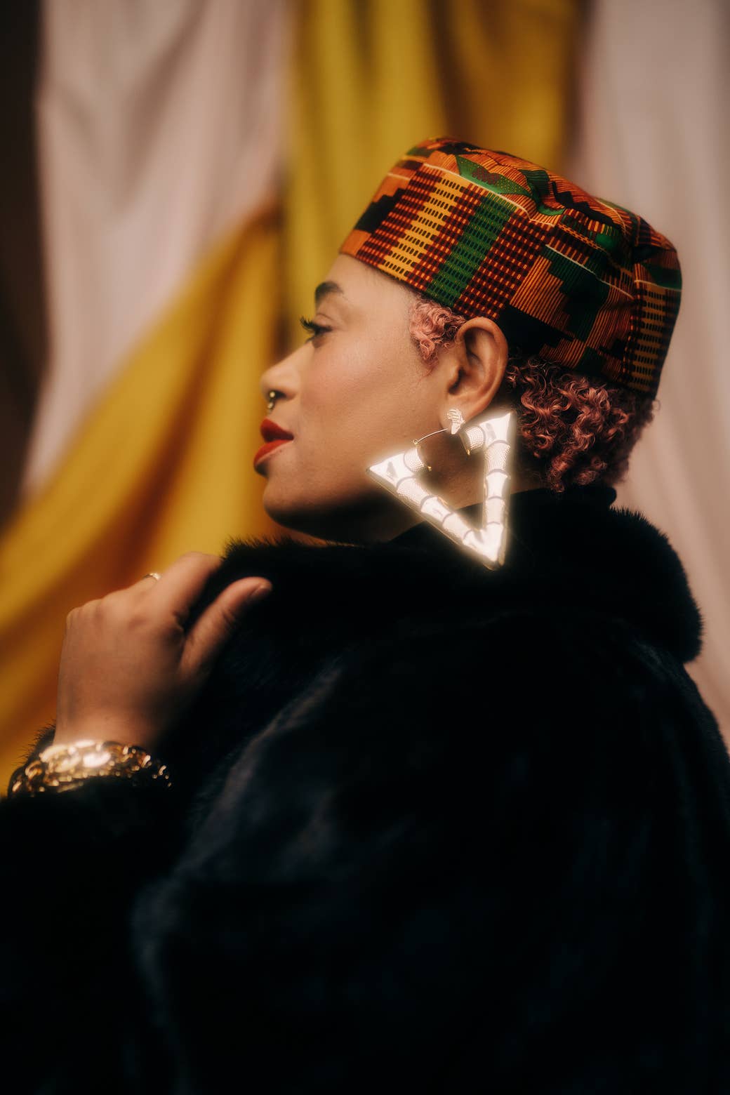 A woman wearing a hat in traditional African print, a black fur coat, and giant gold triangle earrings looks off in the distance.