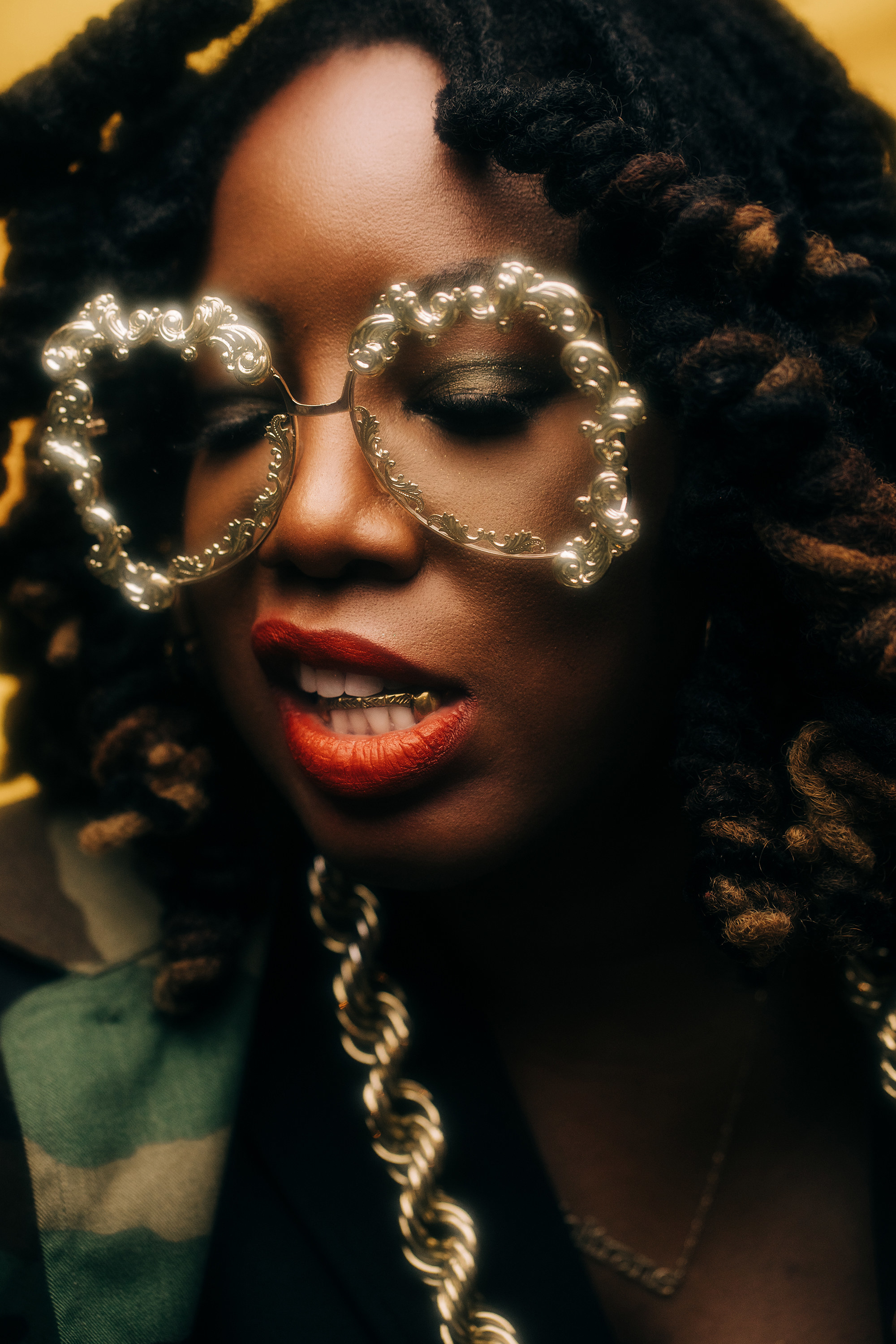 A woman with a god bar in her teeth and giant gold filigree glasses and red liptsick looks down in a portrait.