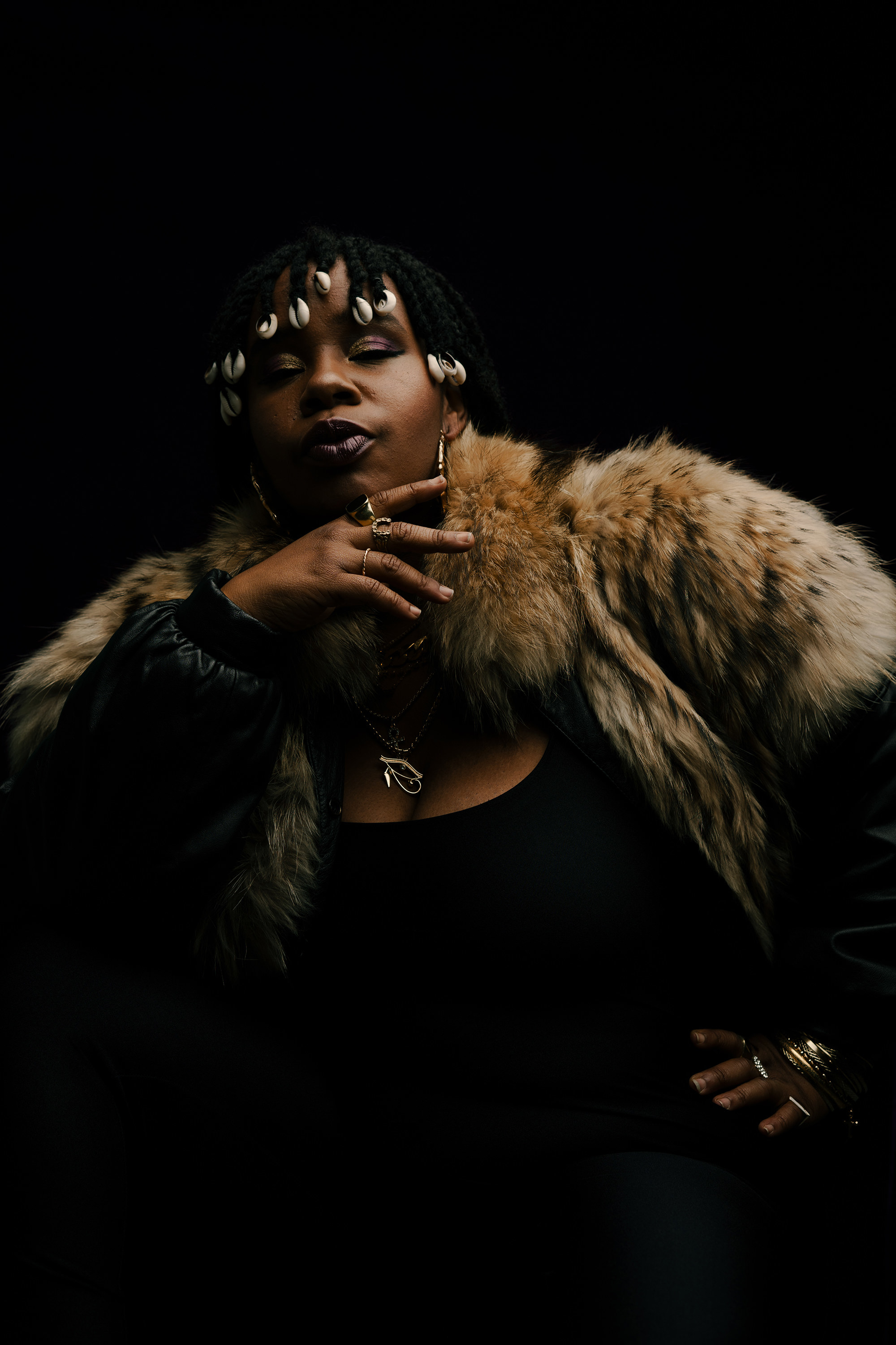 A woman in a brown fur coat, black tank top and twisted braids ended in cowrie shells looks at the camera with her hand to her face.