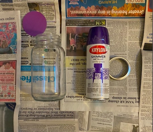 A clear mason jar, a can of spray paint, and a circle cutout are laid out on sheets of newspaper. 