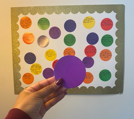 A manicured hand is holding up a paper cutout of a circle in front of a large piece of paper on the wall with quotes. 