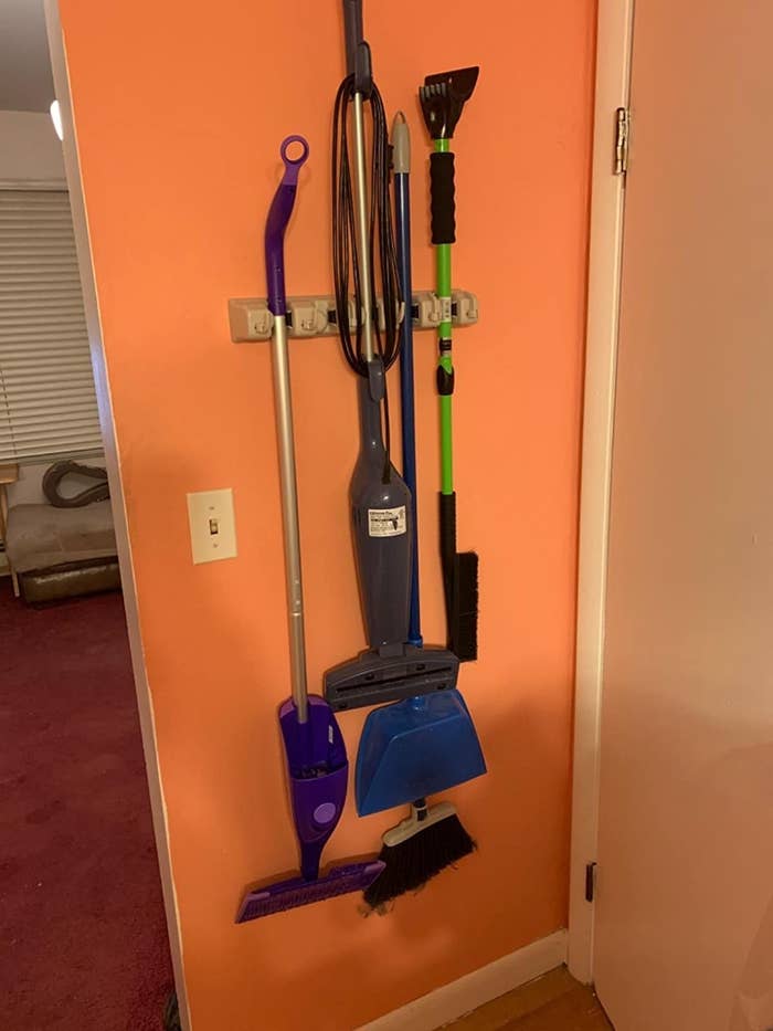a reviewer photo of the mop and broom holder hanging against an orange wall that&#x27;s holding a swifter wet jet mop a steamer two brooms and a dust pan