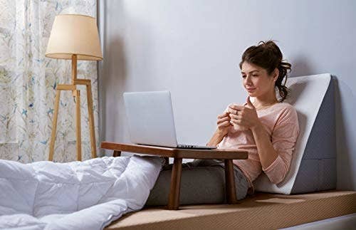 A woman drinking from a mug and watching a laptop in bed, while leaning against an orthopaedic pillow