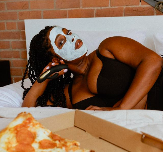 Person in bra and panties laying beside a box of pizza after having applied a face mask. The vibe is in their hand. 