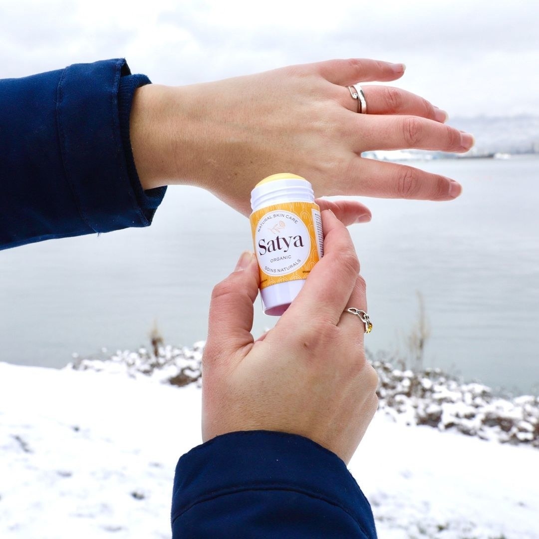 A person applying the lotion stick to their hands in a snowy landscape